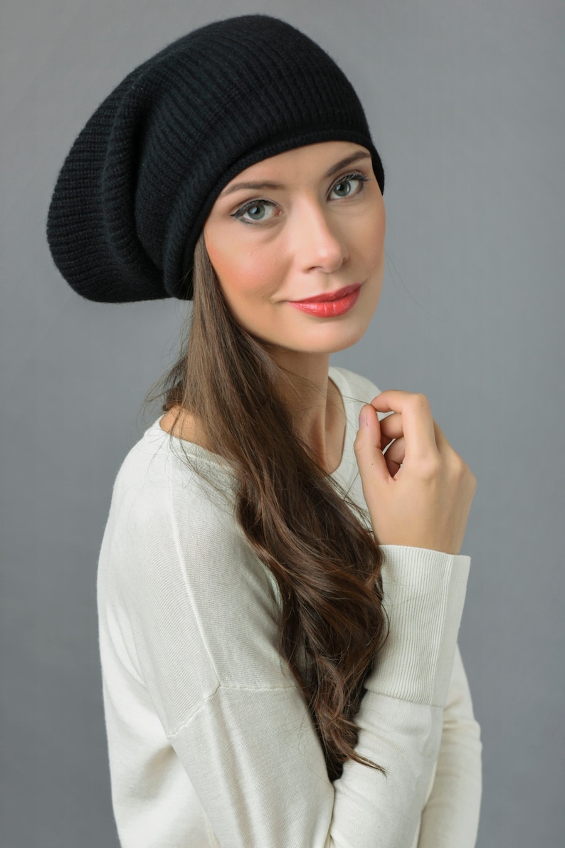 May include: A woman with long brown hair is wearing a black slouchy beanie and a white sweater. She is looking at the camera and smiling.