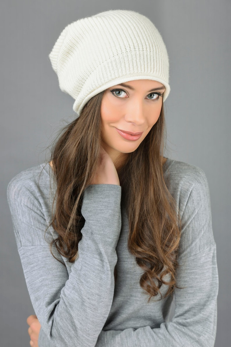 May include: A woman with long brown hair is wearing a white knit beanie and a gray sweater. She is looking at the camera with a slight smile.