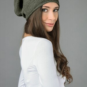 May include: A woman wearing a dark green slouchy beanie hat with a ribbed texture. She is wearing a white long-sleeved shirt.