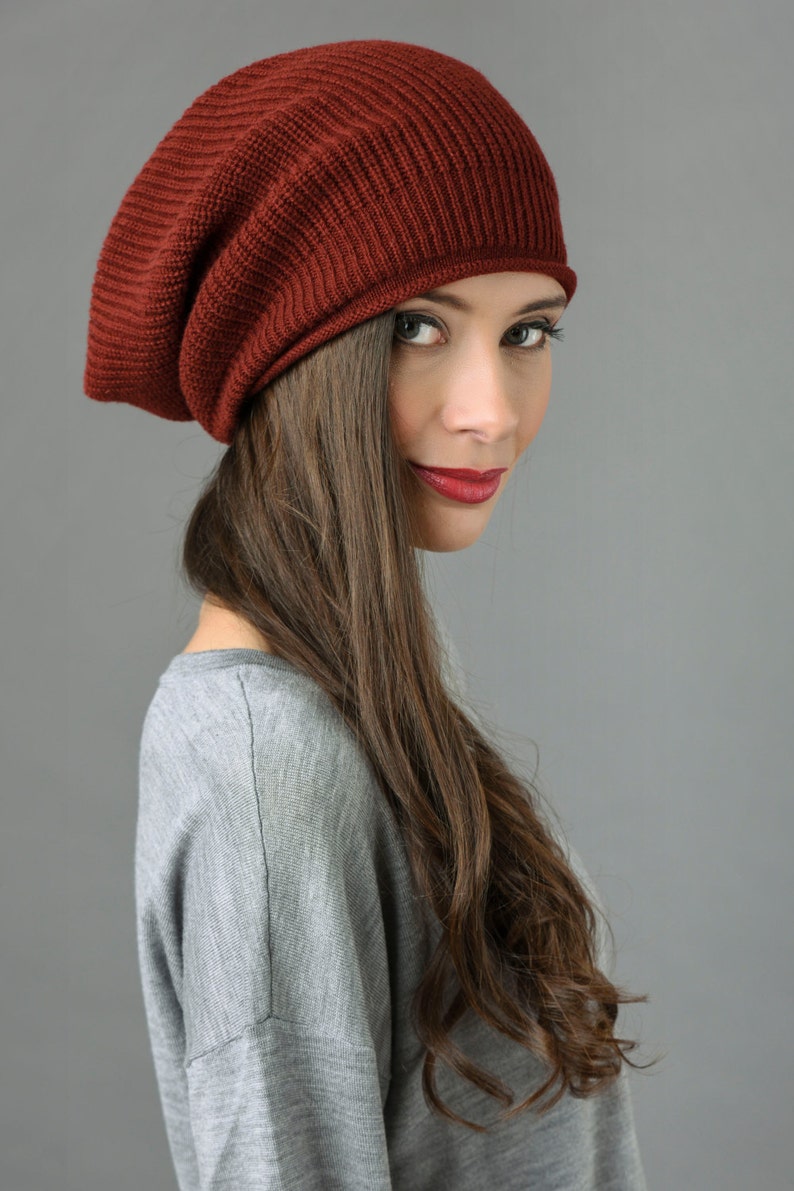 May include: A woman with long brown hair is wearing a slouchy, burgundy beanie. She is looking over her shoulder and smiling.
