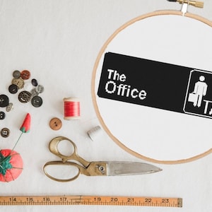 May include: A white embroidery hoop with a black and white cross-stitch design of the "The Office" logo. The logo features the words "The Office" and a silhouette of two people in an office setting. The hoop is surrounded by sewing supplies, including buttons, thread, scissors, and a pincushion.