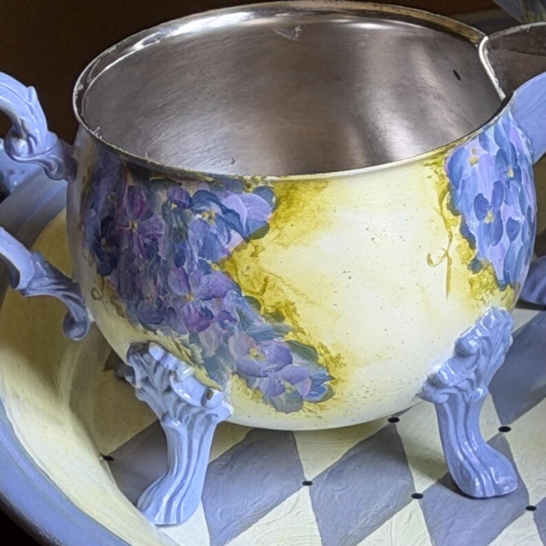 Puede incluir: Una jarrita de color crema con interior met&aacute;lico, decorada con racimos de flores moradas pintadas a mano y patas ornamentadas de color azul claro. La jarrita tiene un asa curva y un pico, y descansa sobre una bandeja a juego con un patr&oacute;n de rombos grises y blancos.