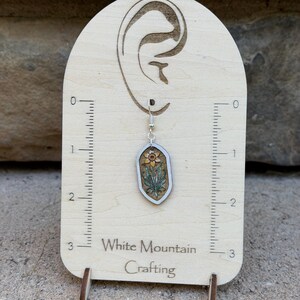 K&ouml;nnte beinhalten: Ein h&auml;ngender Ohrring aus Holz mit einer lasergravierten Narzisse, gr&uuml;nen Bl&auml;ttern und einer gelben Bl&uuml;te, befestigt an einem silberfarbenen Haken auf einer Karte mit 7,6 cm Skala und dem Schriftzug White Mountain Crafting.