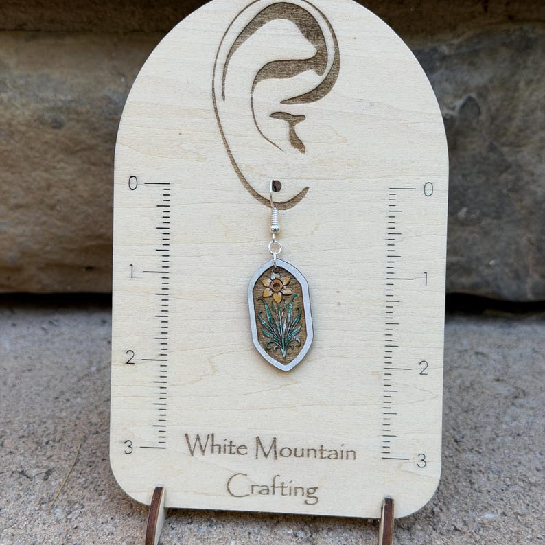 K&ouml;nnte beinhalten: Ein h&auml;ngender Ohrring aus Holz mit einer lasergravierten Narzisse, gr&uuml;nen Bl&auml;ttern und einer gelben Bl&uuml;te, befestigt an einem silberfarbenen Haken auf einer Karte mit 7,6 cm Skala und dem Schriftzug White Mountain Crafting.