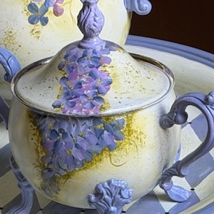 Puede incluir: Un azucarero de color crema con tapa, decorado con flores de glicinia moradas pintadas a mano y motas doradas. El cuenco tiene asas y pies de color morado claro ornamentados, con un remate morado a juego en la tapa.