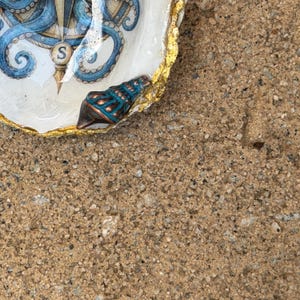 May include: A decorative oyster shell dish featuring a blue octopus and compass illustration with the letter S. The shell has a glossy finish, a gold-painted scalloped edge, and a small, textured, dark teal and copper-colored seashell attached.