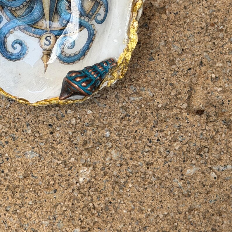 May include: A decorative oyster shell dish featuring a blue octopus and compass illustration with the letter S. The shell has a glossy finish, a gold-painted scalloped edge, and a small, textured, dark teal and copper-colored seashell attached.