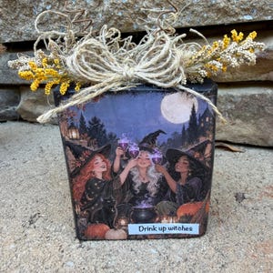 May include: A square wooden block decoration featuring an illustration of three witches toasting with purple drinks, a black cat, pumpkins, and a full moon. The text "Drink up witches" is printed on the front, topped with a natural jute twine bow.