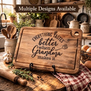 May include: A rectangular wooden cutting board with a juice groove and a corner hole. The board is engraved with the text "Everything tastes better when Grandma makes it" and a small spatula icon. The wood has a natural grain and brown finish.