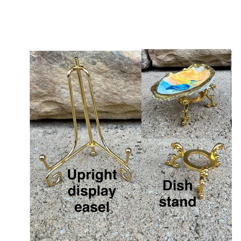 May include: A gold-toned metal upright display easel with curved feet and a gold-toned metal dish stand with three ornate legs, shown supporting a small, irregularly shaped decorative dish with a painted blue, orange, and white interior.