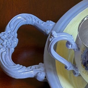 Puede incluir: Una bandeja de servicio de color azul b&iacute;garo claro con asas ornamentadas de volutas en relieve y un centro amarillo p&aacute;lido. La bandeja presenta un acabado pintado liso con intrincados detalles florales en las asas curvas y un borde ancho.
