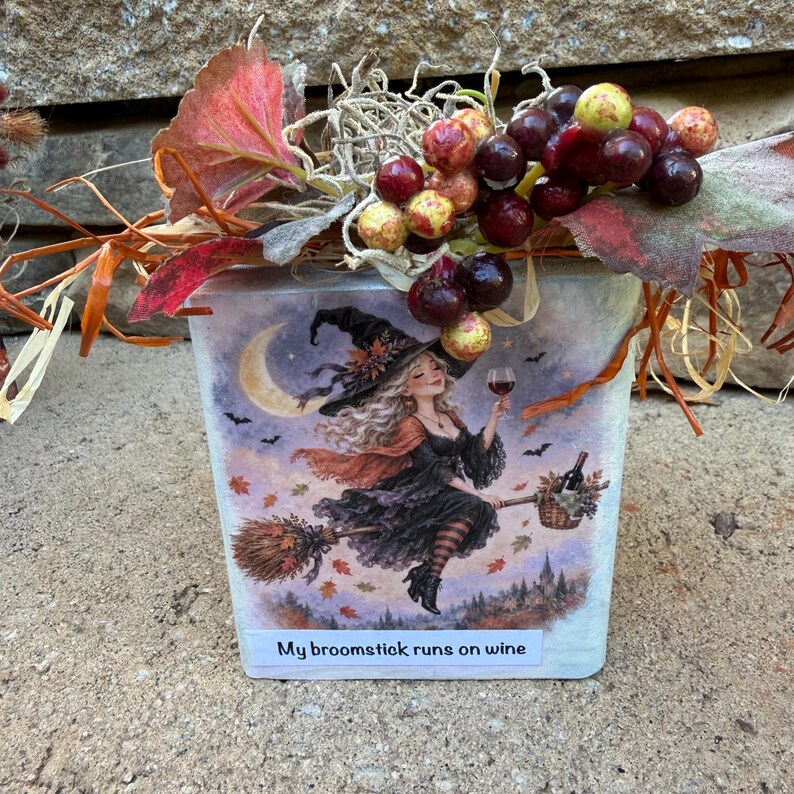 May include: A square decorative block featuring a witch flying on a broomstick with a glass of wine, a crescent moon, and bats. The text reads, "My broomstick runs on wine." The top is adorned with artificial red and purple grapes and autumn leaves.