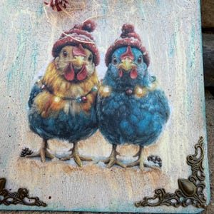 K&ouml;nnte beinhalten: Dekorative Holztafel mit zwei H&uuml;hnern, die rote Strickm&uuml;tzen und Lichterketten tragen. Ein Huhn ist gelb und blau, das andere ist blau. Die Tafel hat dekorative Metallecken und einen strukturierten, verwitterten Hintergrund.