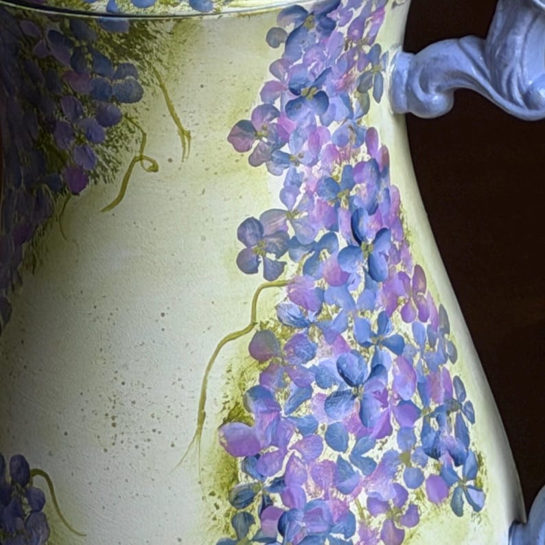 Puede incluir: Una jarra de cer&aacute;mica con un dise&ntilde;o floral pintado a mano que muestra racimos de flores moradas y azules sobre un fondo crema p&aacute;lido. El recipiente incluye un asa curva de color azul claro y delicados detalles de enredaderas verdes.