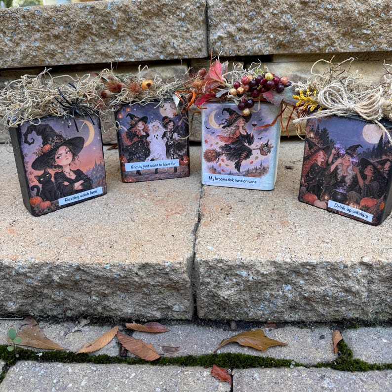 May include: Four square wooden blocks decorated with Halloween-themed illustrations of witches, ghosts, and cats. Each block features text: Resting witch face, Ghouls just want to have fun, My broomstick runs on wine, and Drink up witches, topped with dried moss.