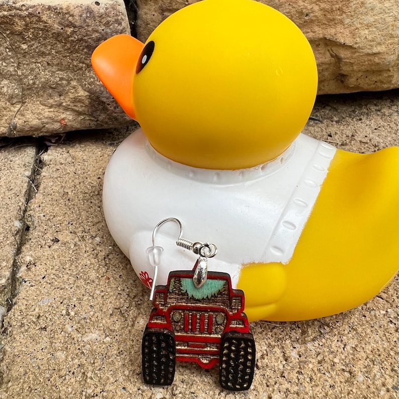 K&ouml;nnte beinhalten: Ein h&auml;ngender Ohrring mit einem roten Gel&auml;ndewagen-Design, schwarzen Reifen und einem t&uuml;rkisfarbenen Akzent auf der Motorhaube. Der Ohrring ist an einem silberfarbenen Metallhaken mit einem durchsichtigen Kunststoffverschluss befestigt.