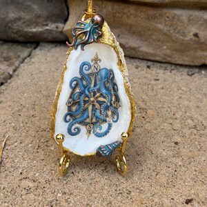 May include: A decorative oyster shell featuring a blue octopus and compass design with the letters N, S, E, and W. The shell has a gold-painted edge, a small metal octopus accent at the top, and a small shell accent at the bottom, displayed on a gold stand.