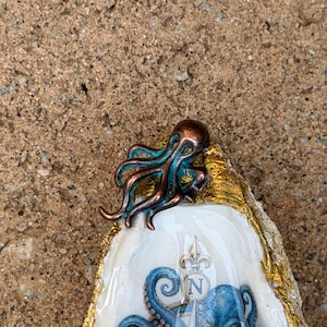 May include: A decorative oyster shell featuring a metallic copper and teal octopus figurine attached to the top edge. The shell has a gold-painted rim and a glossy interior surface displaying a blue octopus illustration and a compass rose with the letter N.
