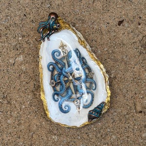 May include: A decorative oyster shell featuring a central compass rose graphic overlaid with a blue octopus illustration. The shell has a gold-painted edge, a small bronze-toned octopus figurine at the top, and a small seashell accent at the bottom.