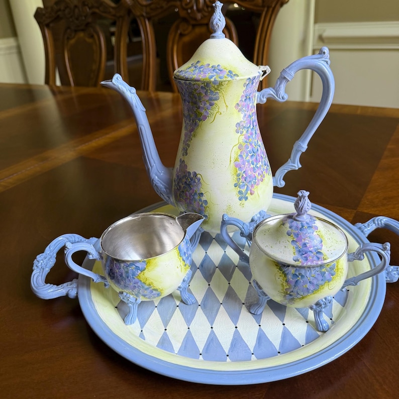Puede incluir: Un juego de t&eacute; que incluye una tetera alta, una jarra de leche y un azucarero con tapa sobre una bandeja redonda. Las piezas est&aacute;n pintadas en amarillo p&aacute;lido con racimos de flores moradas y asas, picos y remates ornamentados en azul claro.