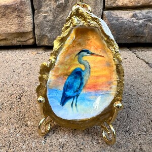 May include: An oyster shell art piece featuring a blue heron painted against a sunset backdrop. The shell is edged with gold paint and mounted on a gold-colored metal stand. The artwork is a decorative coastal-themed item.