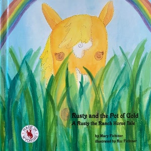 Könnte beinhalten: Kinderbuchcover mit einem gelben Pferd, das durch grünes Gras unter einem Regenbogen blickt. Der Titel lautet "Rusty and the Pot of Gold: A Rusty the Ranch Horse Tale." Das Buch ist "Made in Wyoming."