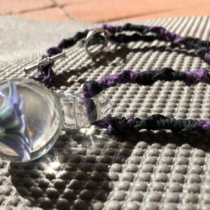 Può includere: Un ciondolo di vetro con un fiore viola all'interno, appeso a un cordoncino intrecciato nero e viola.