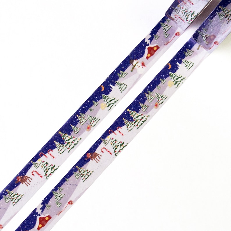 May include: Two rolls of washi tape with a winter forest design. The tape features a snowy landscape with pine trees, a small house, a snowman, and reindeer. The background is a dark blue with white stars.