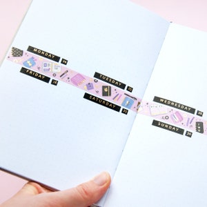 May include: A pink and purple washi tape with gold accents and illustrations of office supplies, including a notebook, pens, and a calculator. The tape is labeled with the days of the week: Monday, Friday, Tuesday, Wednesday, Saturday, and Sunday.