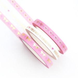 Puede incluir: Rollos de cinta adhesiva decorativa rosa y blanca con detalles dorados. Un rollo presenta el texto "YOU ARE STRONG". Otros rollos tienen corazones y lazos dorados. La cinta es ideal para el scrapbooking y la artesanía.