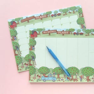 Op de afbeelding: Twee geïllustreerde wekelijkse en maandelijkse plannerpads met een boerderijthema. De pads hebben een groene achtergrond met een rand van bomen, mensen en een rode schuur. Een blauwe pen ligt op de wekelijkse planner. De tekst "PLANS" en "MY WEEKLY PLANS" is zichtbaar.
