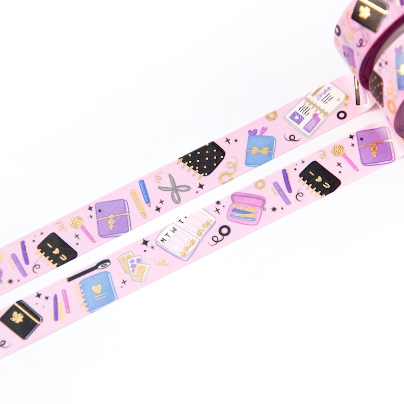 May include: Pink washi tape with a repeating pattern of gold and purple stationery items, including notebooks, pens, pencils, scissors, and a gold glitter star. The tape has the text "MTWTF" and "I love you" on it.