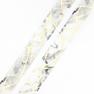 K&ouml;nnte beinhalten: Zwei Rollen wei&szlig;es und goldenes Washi-Tape mit Marmormuster. Das Band hat ein geometrisches Muster aus goldenen Linien auf wei&szlig;em Hintergrund mit grauen Marmorakzenten.