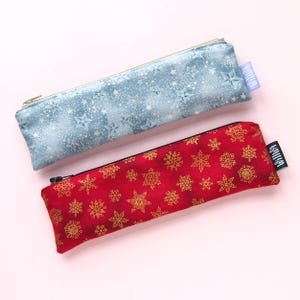 May include: Two zippered pencil cases with a snowflake pattern. One case is blue with white stars and the other is red with gold snowflakes.
