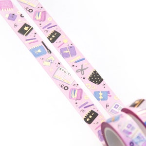 May include: Two rolls of pink washi tape with gold accents. The tape features a repeating pattern of colorful stationery items, including notebooks, pens, pencils, and scissors. The text "MTWT" is visible on one of the notebooks.