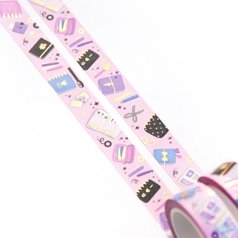 May include: Two rolls of pink washi tape with gold accents. The tape features a repeating pattern of colorful stationery items, including notebooks, pens, pencils, and scissors. The text "MTWT" is visible on one of the notebooks.