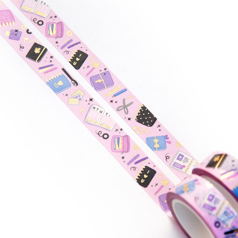 May include: Two rolls of pink washi tape with a gold glitter background and a repeating pattern of colorful illustrations of notebooks, pens, pencils, scissors, and other stationery items. The text "MTWT" and "I love you" are visible on the tape.