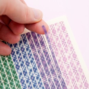 Può includere: Un foglio di nastro washi a motivi con vari disegni a quadri in tonalità di viola, blu e verde. Il nastro viene staccato dal foglio.