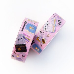 May include: Two rolls of pink washi tape with gold foil accents. The tape features illustrations of notebooks, pens, and other stationery items. The text on the tape reads "MTWThFSS" and "9".