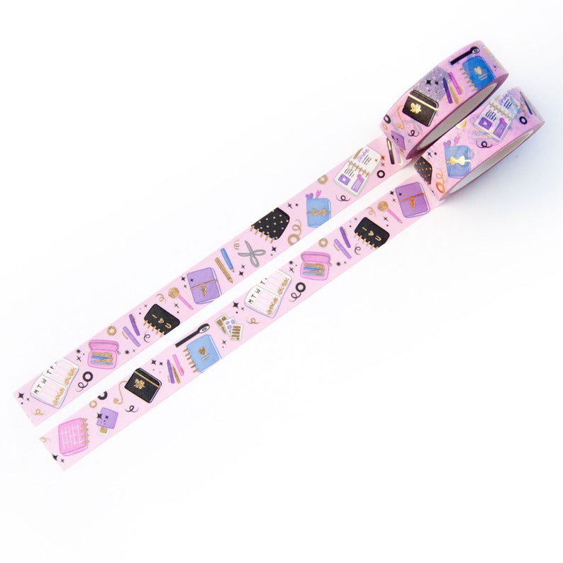 May include: Two rolls of pink washi tape with a gold and purple pattern of notebooks, pens, and other stationery items. The tape is on a white background.