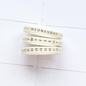 Puede incluir: Tres rollos de cinta washi blanquecina con letras doradas. El rollo superior dice "MIÉRCOLES", el rollo del medio tiene los días de la semana y el rollo inferior muestra los números 20-28. La cinta está apilada sobre una superficie blanca punteada.