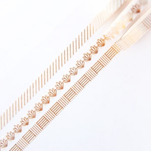 Op de afbeelding: Drie decoratieve washi-tapes met goudfolie-ontwerpen op een witte achtergrond. De tapes hebben verschillende patronen: diagonale lijnen, bloemmotieven en geometrische vormen. De tapes zijn schuin geplaatst en tonen hun ontwerpen.