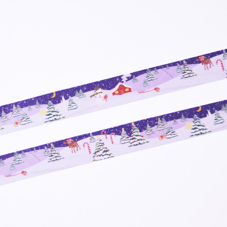May include: Purple and white washi tape with a winter scene featuring snow-covered trees, a red cabin, a reindeer, and candy canes. The tape has a glittery finish.