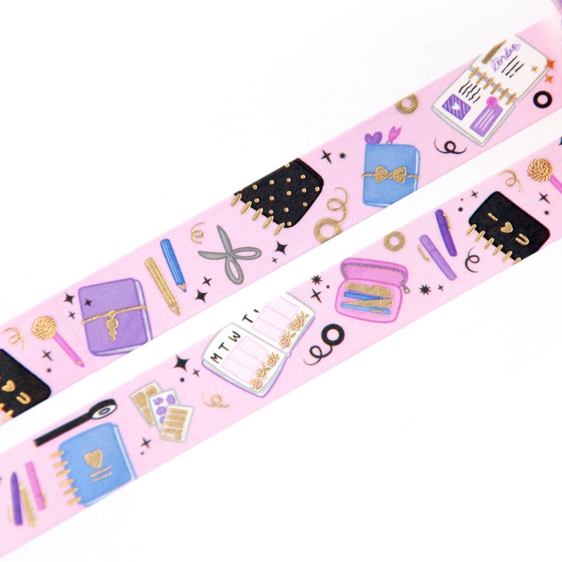 May include: Pink washi tape with a repeating pattern of gold and purple illustrated notebooks, pens, pencils, scissors, and other stationery items. The tape has a gold glitter finish.