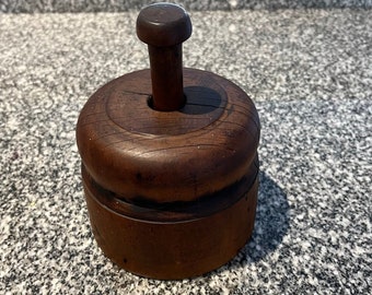 May include: A vintage wooden butter mold with a round base and a wooden knob on top. The mold is dark brown and has a worn, rustic appearance.