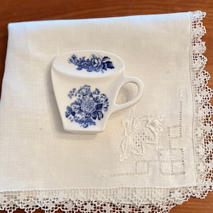 May include: A white ceramic teacup with a blue floral design. The teacup is sitting on a white linen napkin with lace trim.