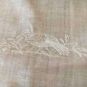 May include: White embroidered bird design on a white linen fabric. The bird is facing right and has a long tail. The design is surrounded by leaves and flowers.