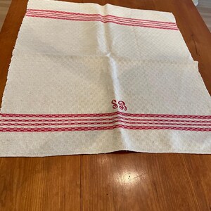 May include: Cream-colored linen towel with red stripes and the initials "SR" embroidered in red. The towel has a textured weave and is folded on a wooden surface. The red stripes are near the top and bottom edges.