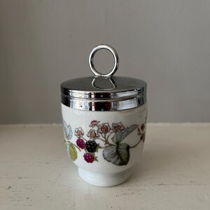 May include: A white ceramic egg cup with a silver lid and a floral design featuring blackberries and leaves. The lid has a silver ring handle.