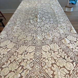 May include: Cream-colored lace tablecloth with a floral pattern. The intricate design features detailed flowers and leaves, creating a delicate and elegant look. The tablecloth is rectangular and covers a wooden table.